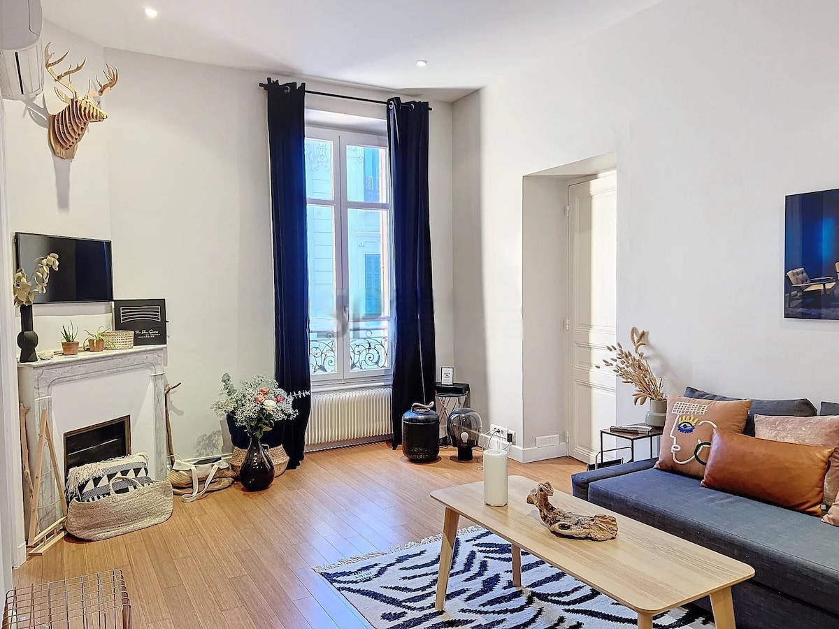 A living room features natural light streaming through tall windows, accompanied by dark curtains. A comfortable sofa is positioned opposite a stylish coffee table. Decorative elements include a fireplace and potted plants, creating a welcoming ambiance. A piece of wall art provides a focal point in the space.