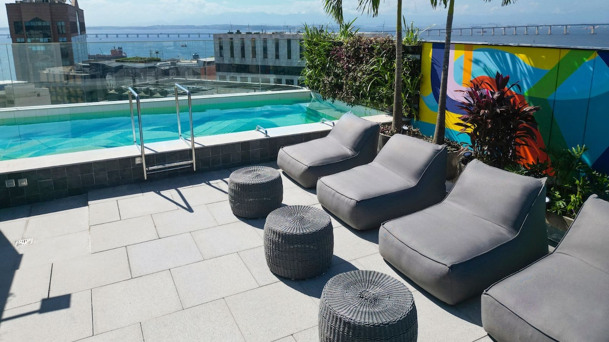 A rooftop pool area is showcased, featuring a clear swimming pool surrounded by stylish lounge chairs. The flooring consists of large tiles, and vibrant greenery and mural enhance the modern ambiance. A serene view of the ocean is visible in the background.