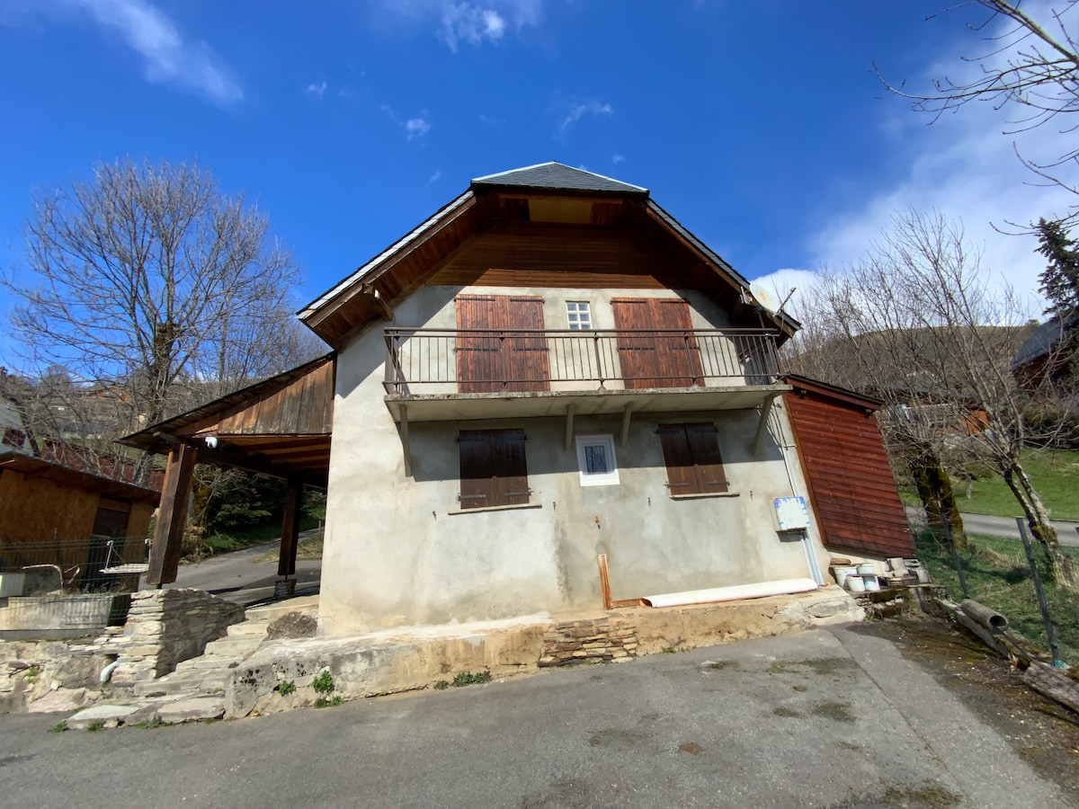 The exterior of the house presents a blend of wooden and contemporary design elements. Balanced between two levels, the structure features a small balcony with shutters and a simple pathway leading to the entrance. Surrounding greenery is visible, enhancing the setting.