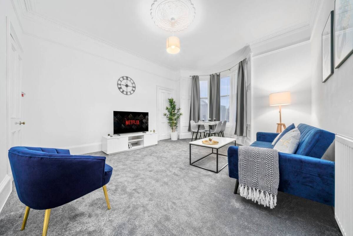 A spacious living area features soft gray carpeting and white walls. A blue armchair and a matching sofa are arranged around a coffee table. A TV sits atop a white media cabinet, while a dining table and chairs are positioned in the background. Natural light filters through large windows, with drapes softly framing them.