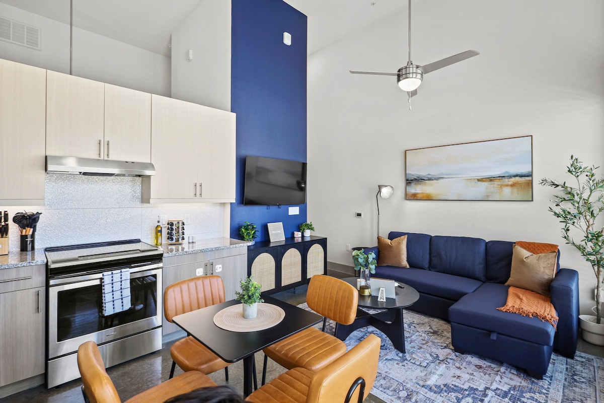 A stylish living area features a comfortable sectional sofa and a sleek coffee table set against a backdrop of light cabinetry. A mounted television and modern ceiling fan enhance the space, while decorative elements, including a potted plant, complete the inviting atmosphere.