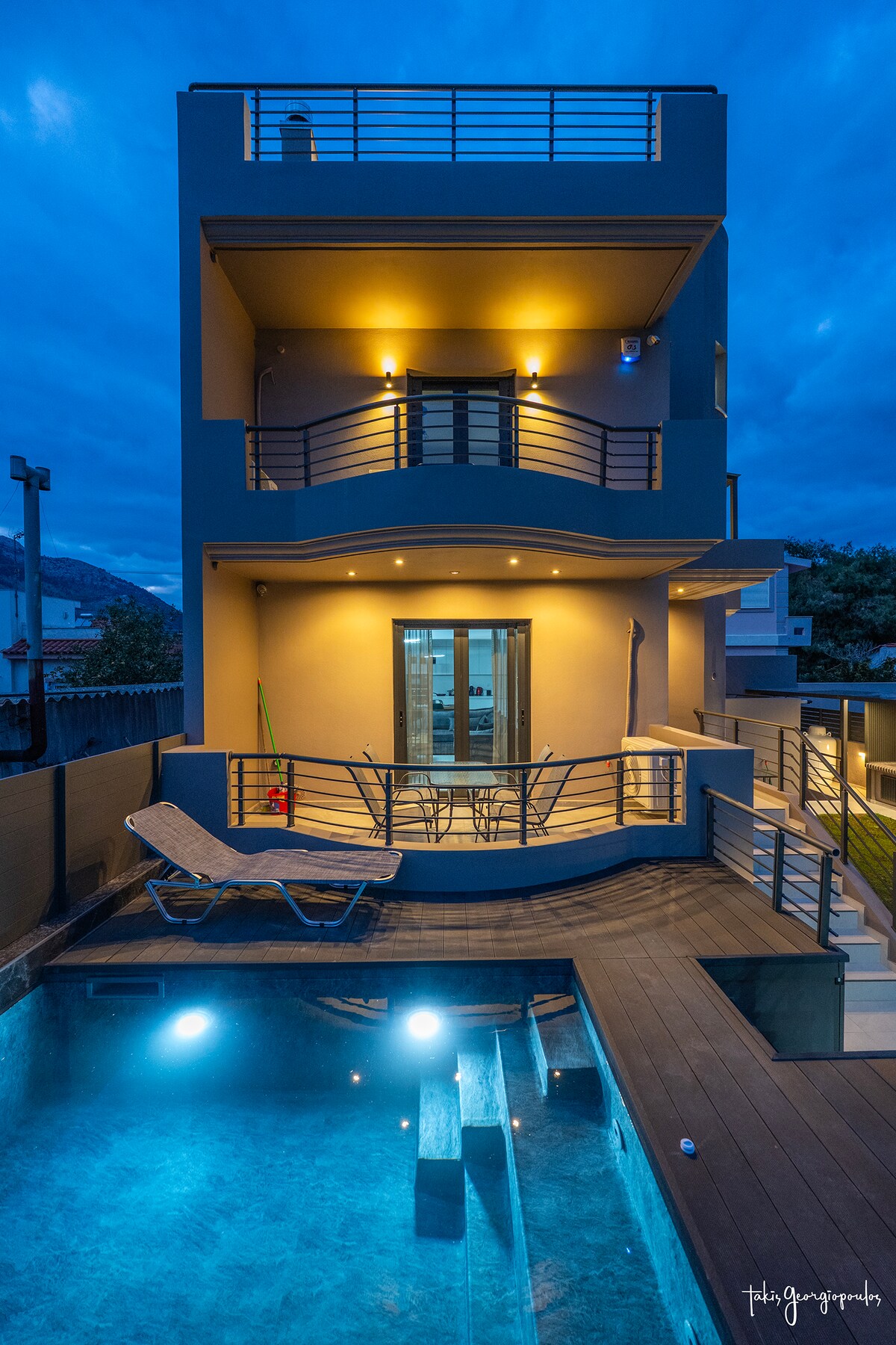 The exterior of the contemporary villa is captured at dusk, highlighting a private pool with illuminated water. Spacious balconies and seating areas are visible, while warm lights enhance the building's structure against a backdrop of a serene evening sky.