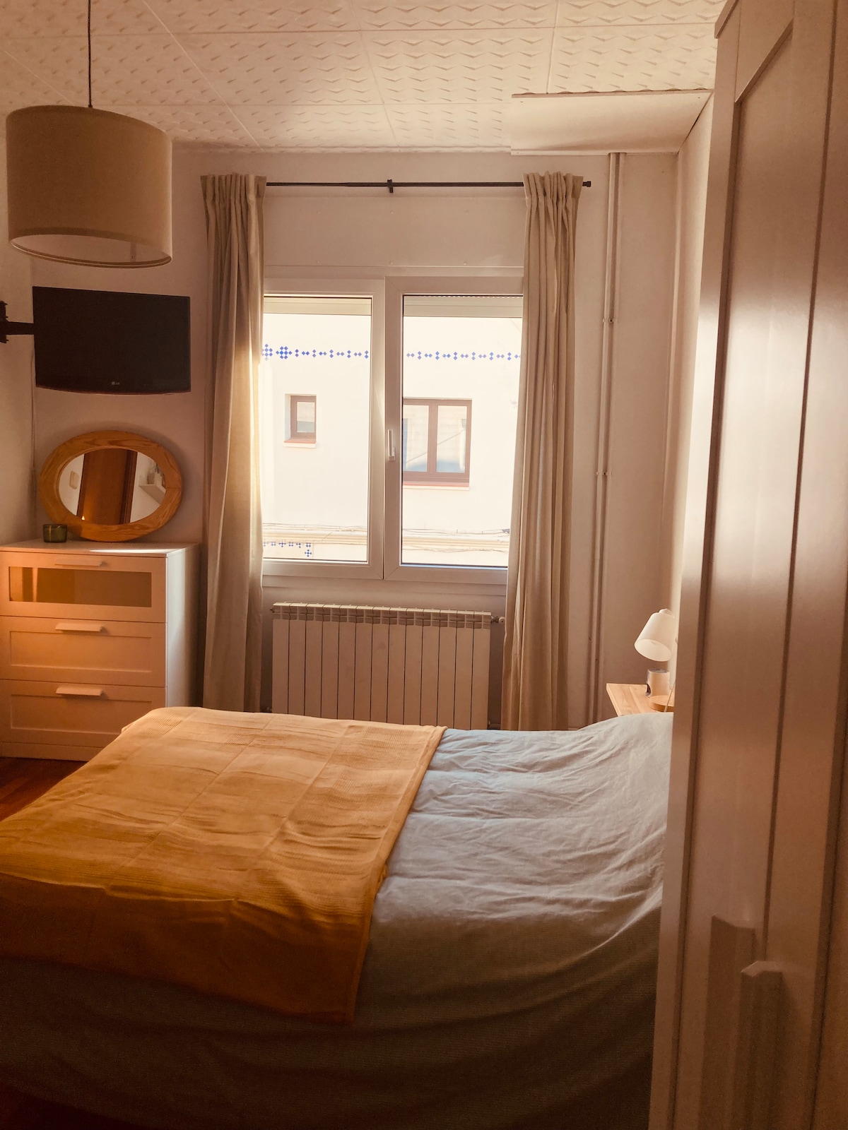 A cozy bedroom is presented with a spacious bed dressed in light bedding. A window allows natural light to enter, complemented by light-colored curtains. A small dresser and a round mirror are visible, adding functionality to the space, along with a wall-mounted television.