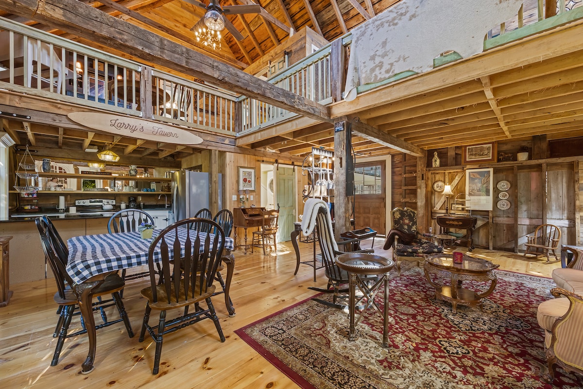 The rustic interior features exposed wooden beams and a warm wooden floor. A large table with black chairs is located in the dining area, complemented by a patterned rug. An inviting seating area includes plush chairs, and a spacious kitchen can be seen in the background.