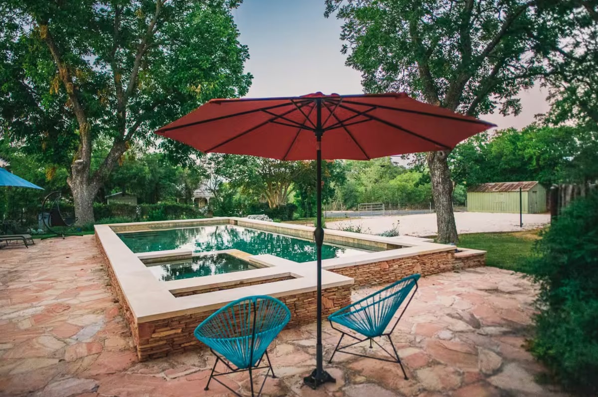 An outdoor area features a stone patio surrounding a rectangular pool, complemented by an umbrella providing shade. Two blue accent chairs with a modern design are positioned near the pool, surrounded by lush greenery and mature trees, creating a serene outdoor setting.