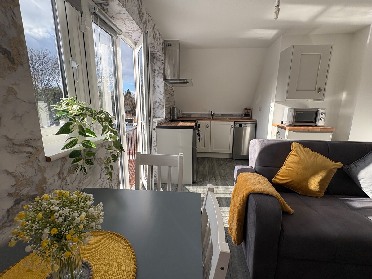 A bright kitchen area features modern appliances and a cozy dining table. Sunlight floods in through large windows, illuminating soft furnishings, including a grey sofa adorned with a mustard throw. A small vase of flowers sits on the dining table, adding a touch of warmth to the space.