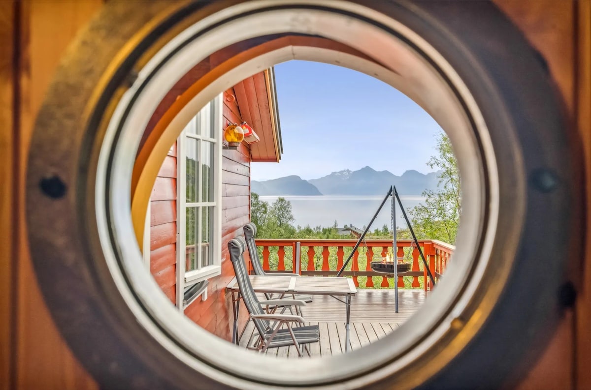 A circular window frame offers a view of a serene landscape, featuring distant mountains and a calm body of water. The foreground includes a spacious deck with seating, framed by a vibrant red wooden cabin.