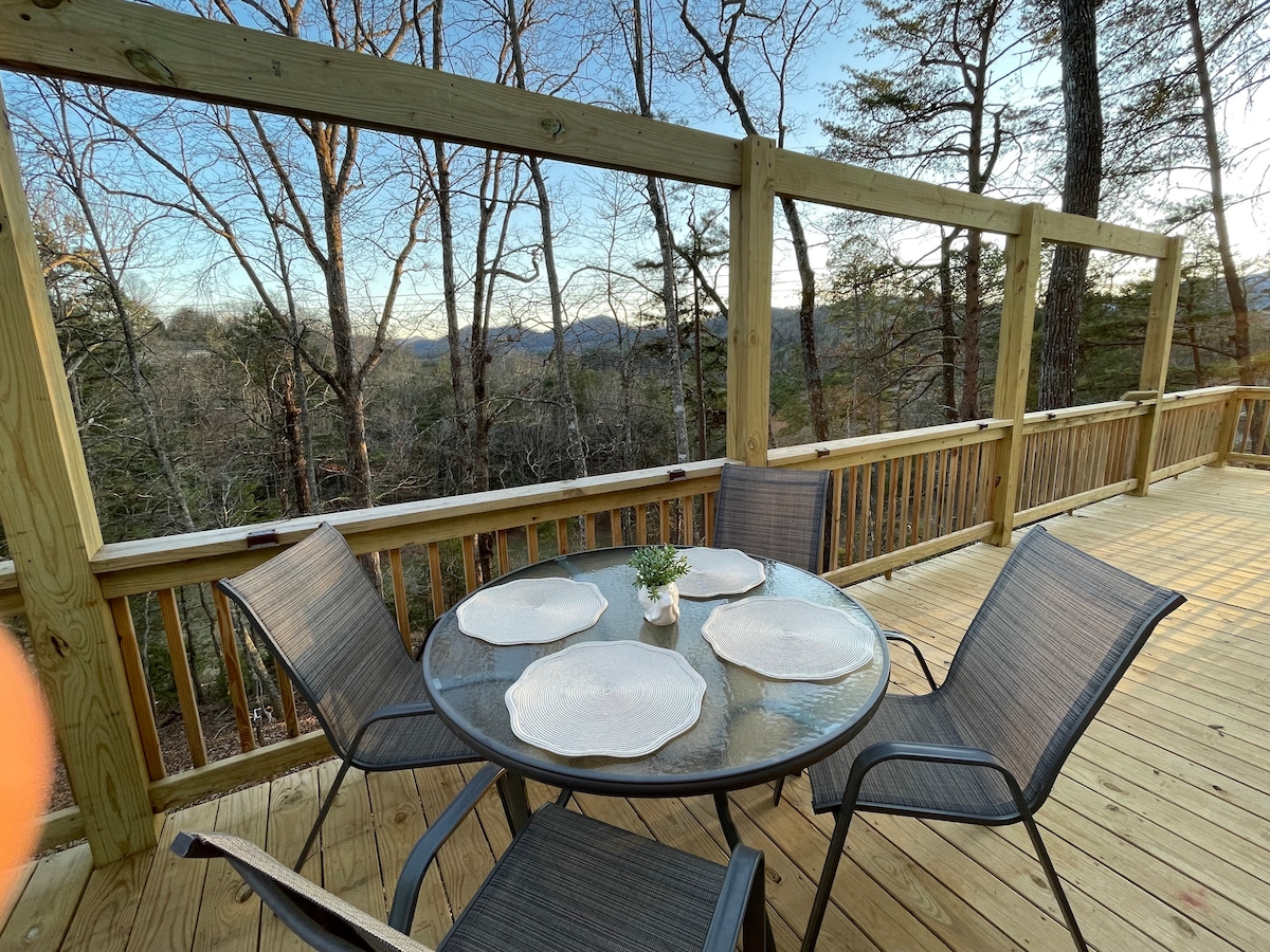 A round table is set for dining on a wooden deck, surrounded by four chairs. Mountain views are visible beyond a railing, featuring trees in the foreground and distant peaks under a clear sky.