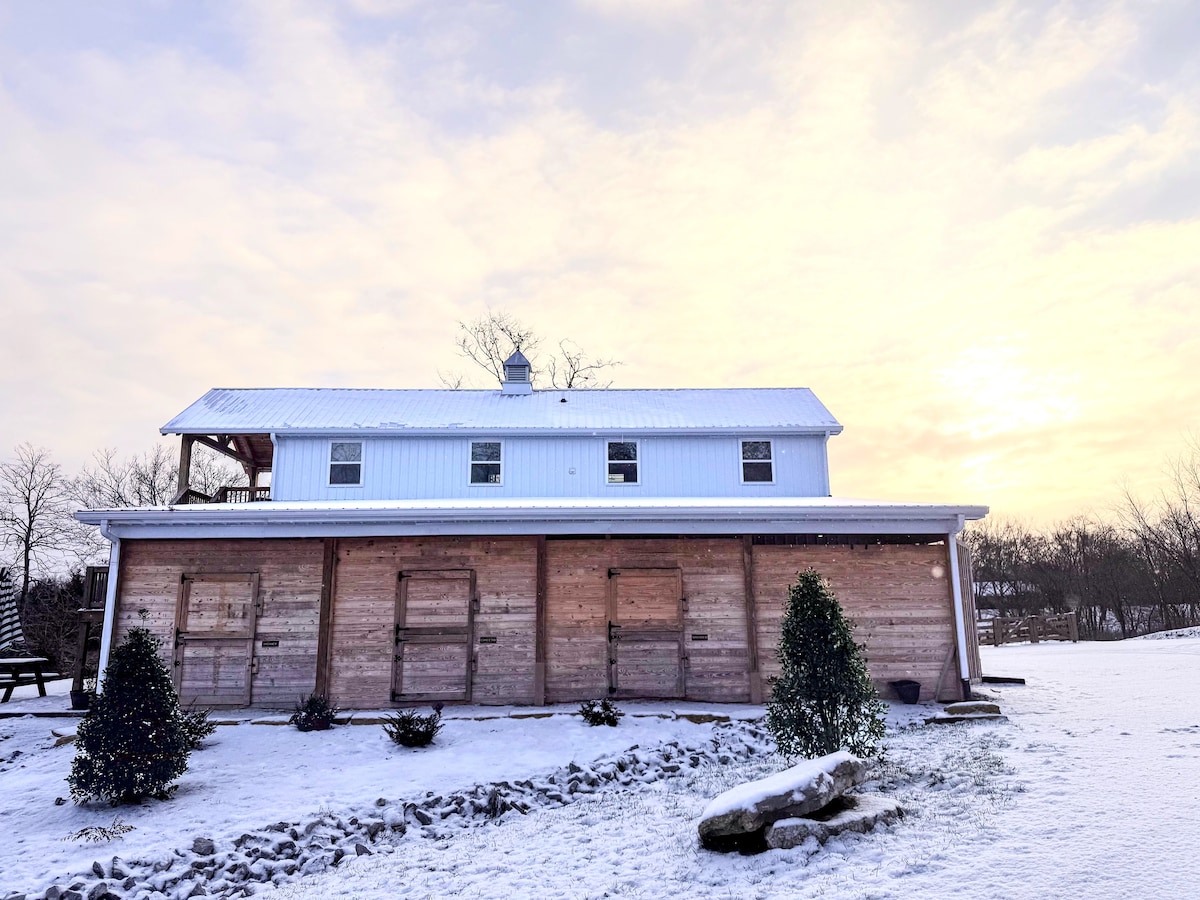The exterior of the luxury barn apartment is visible, showcasing a clean, wooden structure illuminated by soft sunlight under a cloudy sky. A dusting of snow covers the ground, highlighting the building's warmth and rustic style. Neatly trimmed shrubs and a stone pathway accentuate the entrance.