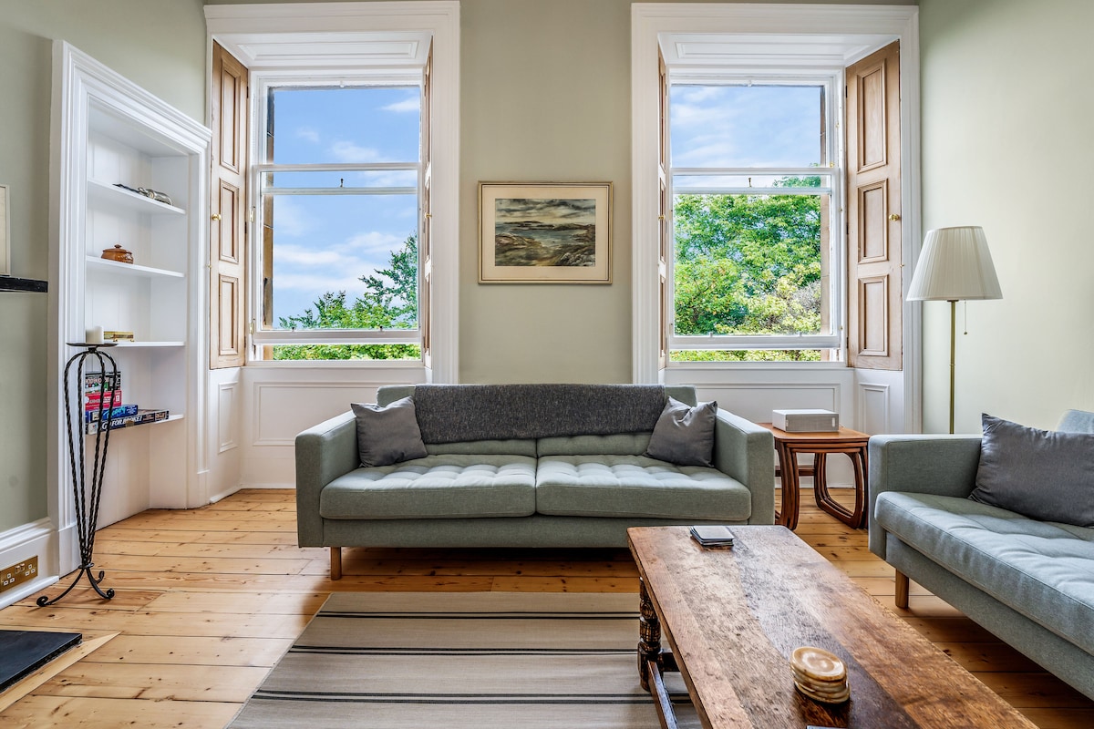 A stylish living room features large windows with traditional shutters that open to greenery outside. A comfortable sofa with a light grey hue is paired with a wooden coffee table. Natural light fills the space, highlighting the light wooden flooring and gentle colour scheme.