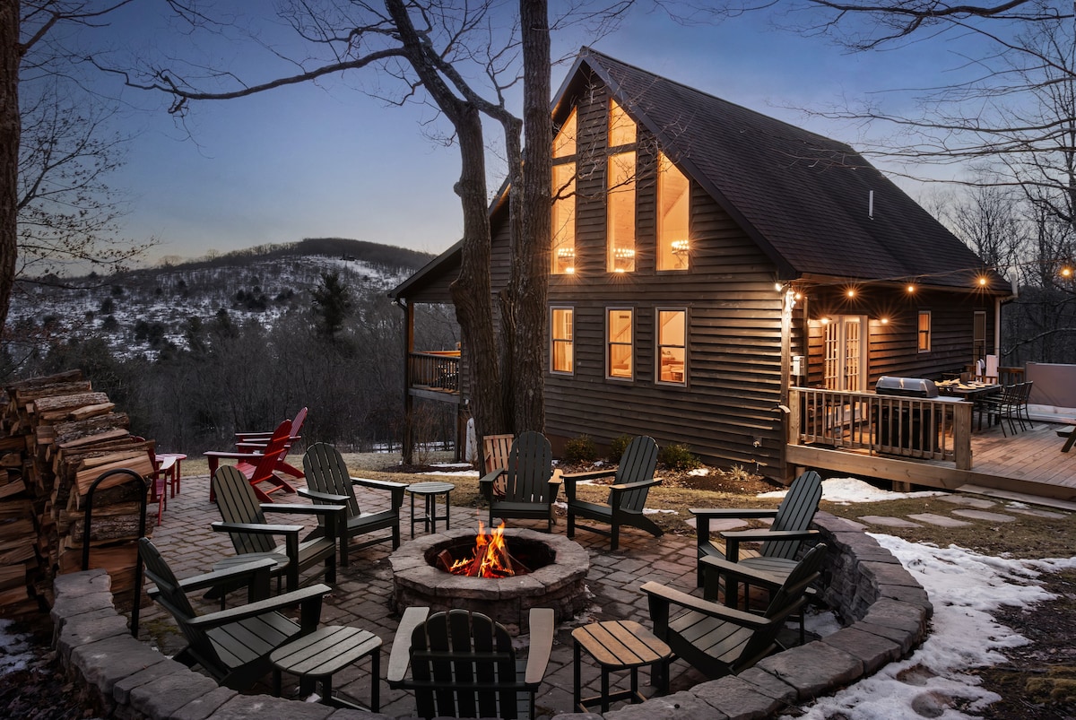 A spacious cabin exterior is showcased, featuring large windows and wooden siding. A stone fire pit is surrounded by multiple Adirondack chairs, inviting gatherings. Nearby, stacks of firewood and a deck with outdoor seating are visible, all set against a serene forest landscape at dusk.