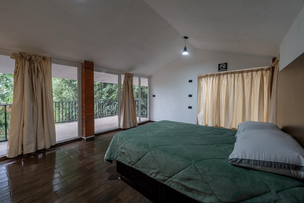 A spacious sleeping area features a large bed with a green blanket and two pillows. Natural light enters through large windows adorned with curtains, offering views of the surrounding greenery. The room includes a warm wood floor and a soft color palette.