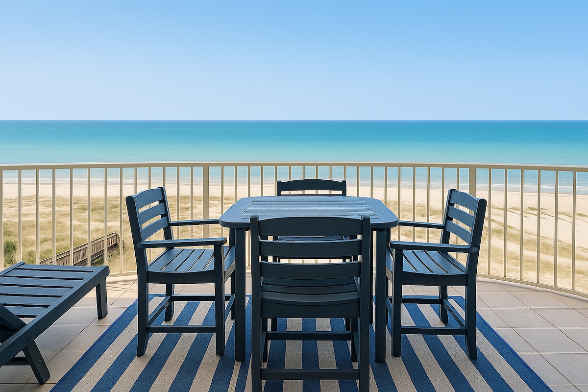 A private balcony is furnished with a bistro dining set and several lounge chairs, all overlooking a vibrant coastline. The view includes a clear blue sky meeting the tranquil sea, enhancing the serene coastal atmosphere.