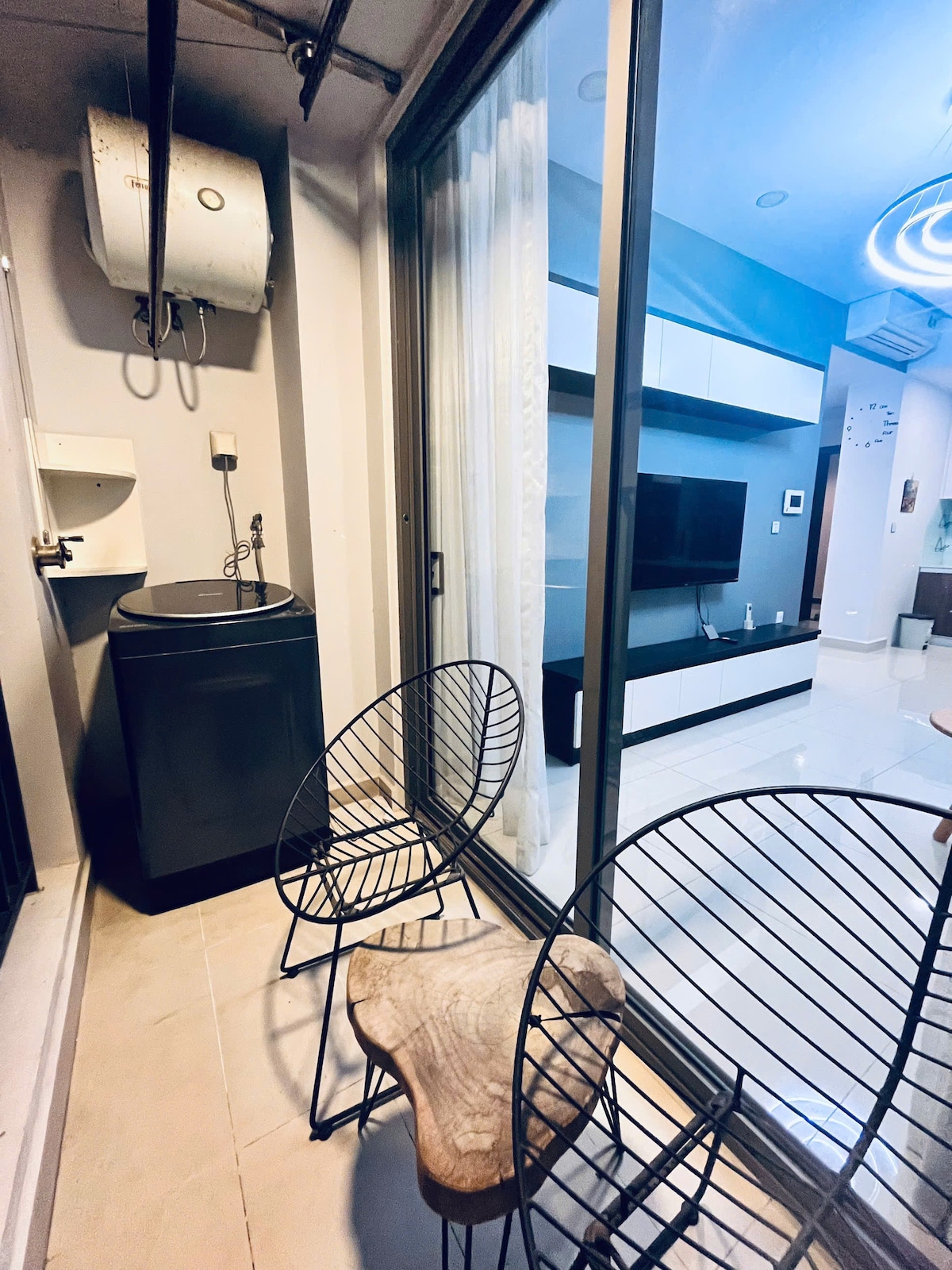 A small balcony area features two wire-framed chairs placed next to a round wooden table. A washing machine is positioned against the back wall, while large glass doors lead into the living space, allowing natural light to enter.