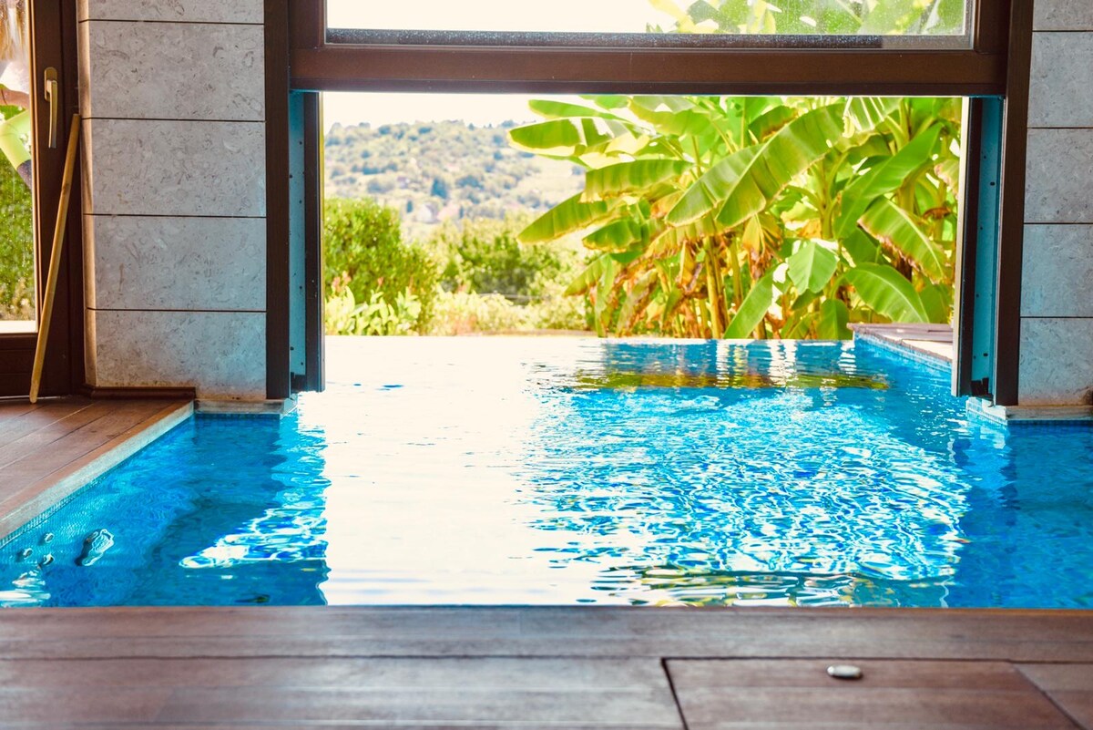 The image captures a serene infinity pool, seamlessly extending into a lush garden view. Clear blue water reflects the sunlight, inviting relaxation. Framing the pool are large glass doors that open onto a vibrant outdoor space featuring tropical greenery.