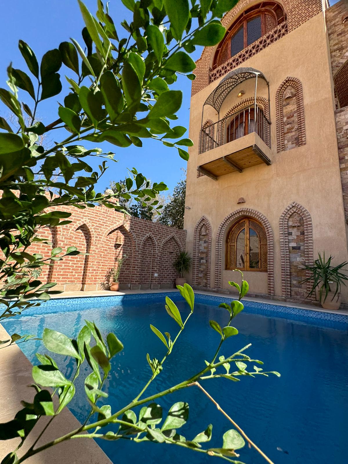 A tranquil garden view is captured with lush greenery framing a private swimming pool. The villa's architectural details, including arched windows and textured stone walls, are visible in the background, adding to the serene ambiance of the outdoor space.