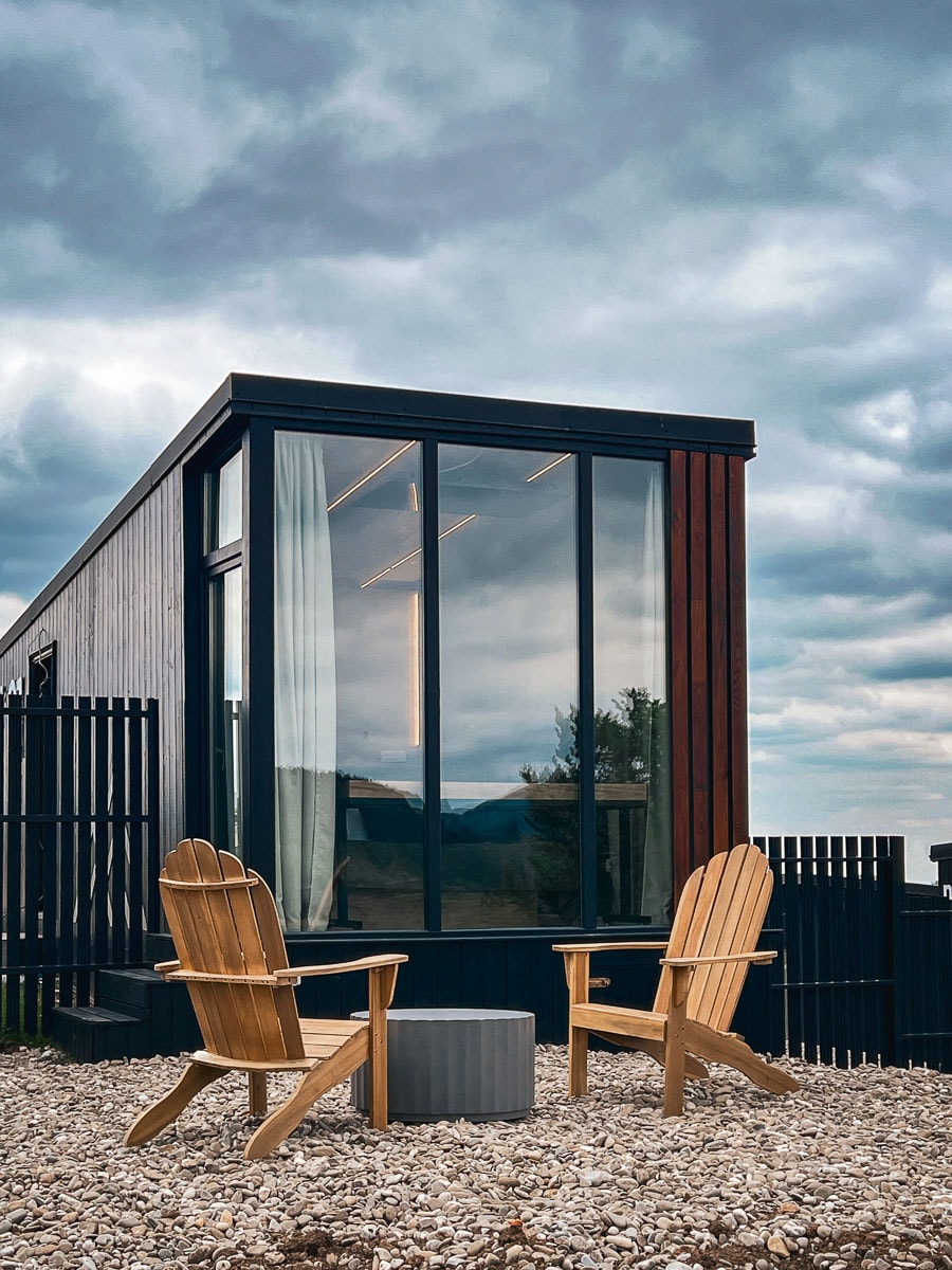 The exterior of the cabin features large windows framed in black, allowing for natural light to flood the interior. Two wooden Adirondack chairs are positioned in front of a small, round table, resting on a stone surface, with a backdrop of a cloudy sky.