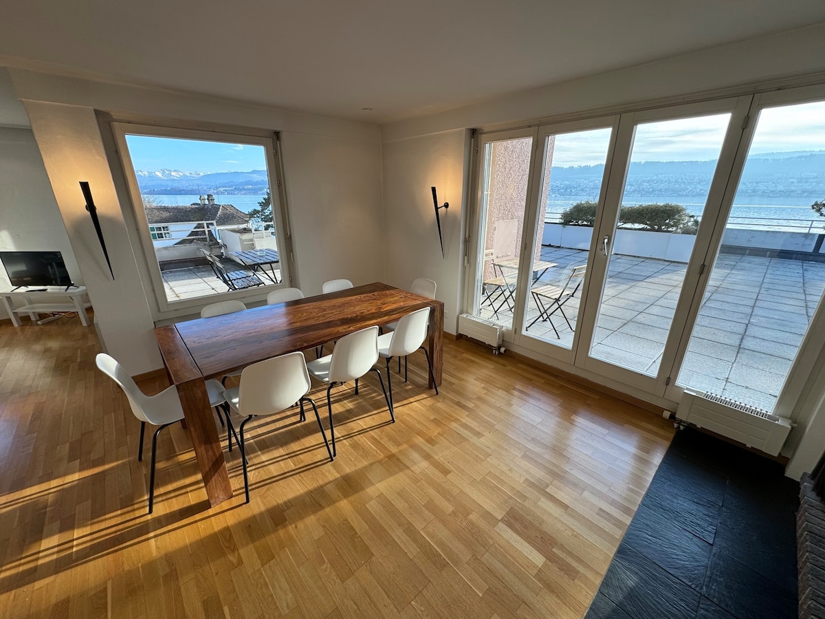 A dining area is presented with a wooden table and six white chairs. Large glass doors provide ample natural light and reveal a scenic terrace overlooking Lake Zurich. A view of the mountains in the distance enhances the serene ambiance.