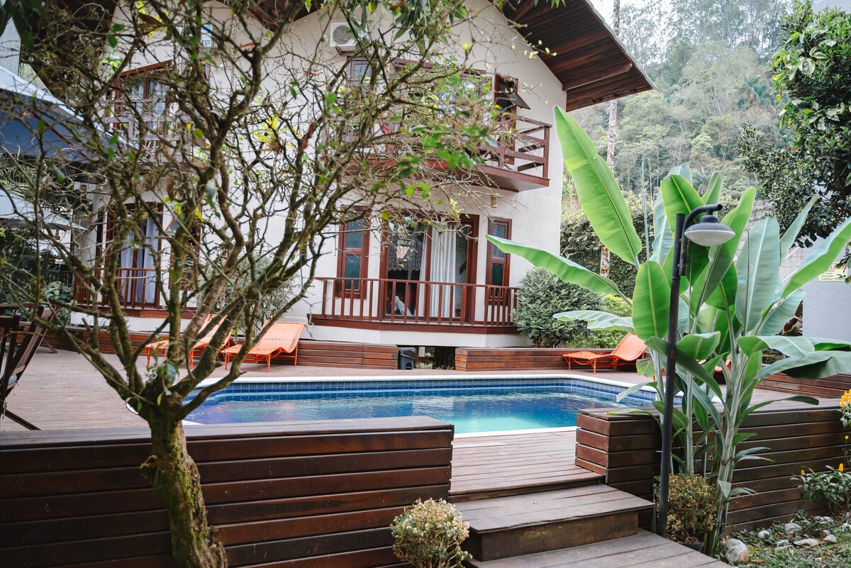 A two-story house is framed by trees and tropical plants, featuring a wooden deck surrounding a rectangular swimming pool. Lounge chairs in bright orange are positioned by the pool, and large glass doors offer views into the inviting interior space.