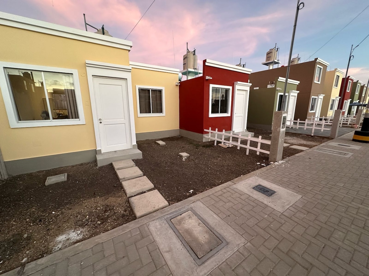 A row of colorful houses is seen, featuring a variety of exterior colors including yellow, red, and green. Each unit has a door and a window, accompanied by a simple white picket fence along the grassy front yard. A brick pathway leads to the doors.