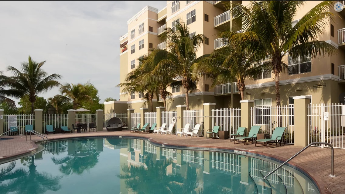 A spacious outdoor pool is surrounded by lounge chairs and palm trees, reflecting the building's facade. The area is enclosed with a white fence, providing a sense of privacy. Barbecue facilities are available nearby, inviting outdoor gatherings.