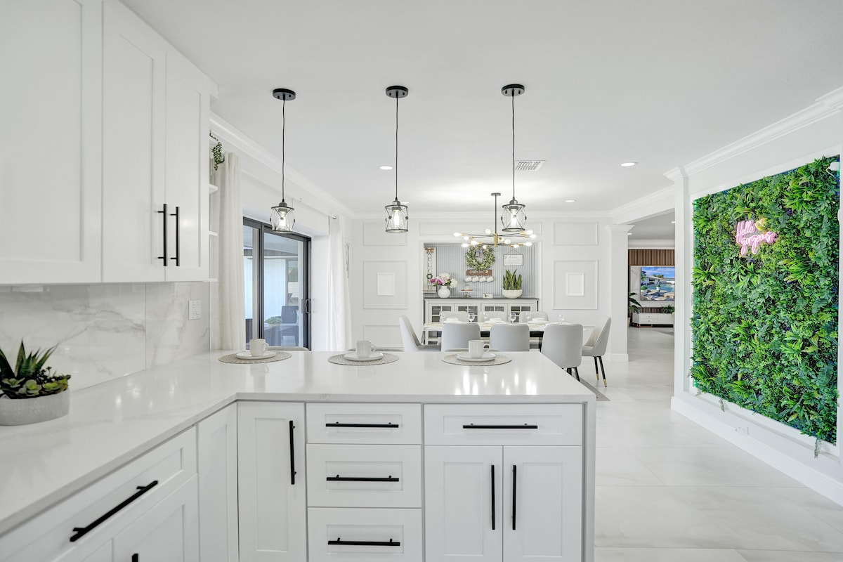 A modern kitchen is available, featuring white cabinetry and sleek black hardware. Pendant lights hang above the island, which showcases a minimalist design. Light streams in from windows adjacent to the dining area, complemented by greenery on a wall and a tasteful decor arrangement.