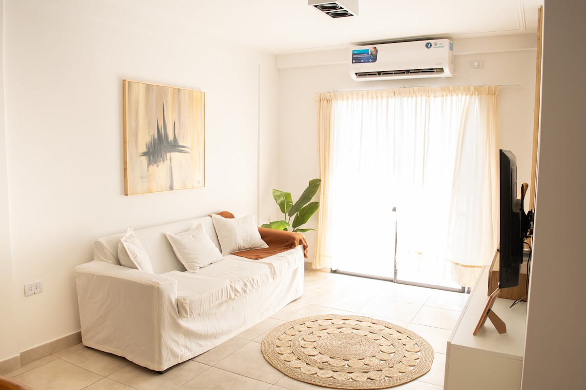 A well-lit living area features a white sofa in front of a large window with light curtains. A round woven rug adds texture to the tiled floor. A green plant is positioned beside the window, while an air conditioning unit is mounted on the wall.