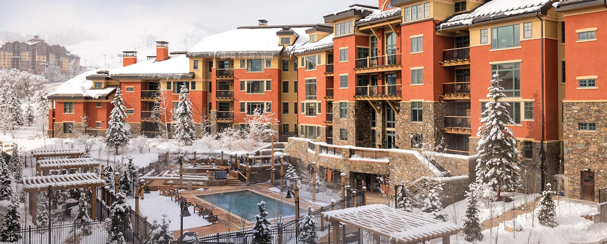 The resort is showcased in a winter landscape, surrounded by snow-covered trees. The architecture features a mix of colorful facades and stone elements. An outdoor swimming pool is visible, bordered by decorative structures, enhancing the winter ambiance.