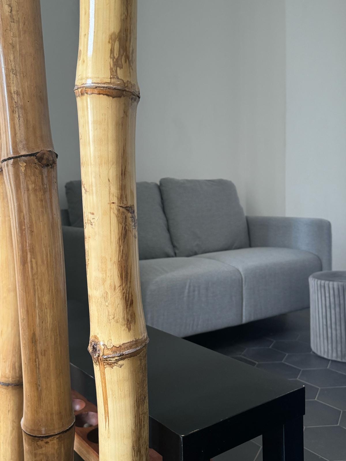 A comfortable gray sofa is positioned against a neutral wall, partially obscured by bamboo accents. A small ribbed stool complements the seating area, creating a cozy corner within the space. The flooring features dark tiles, adding to the overall ambiance.