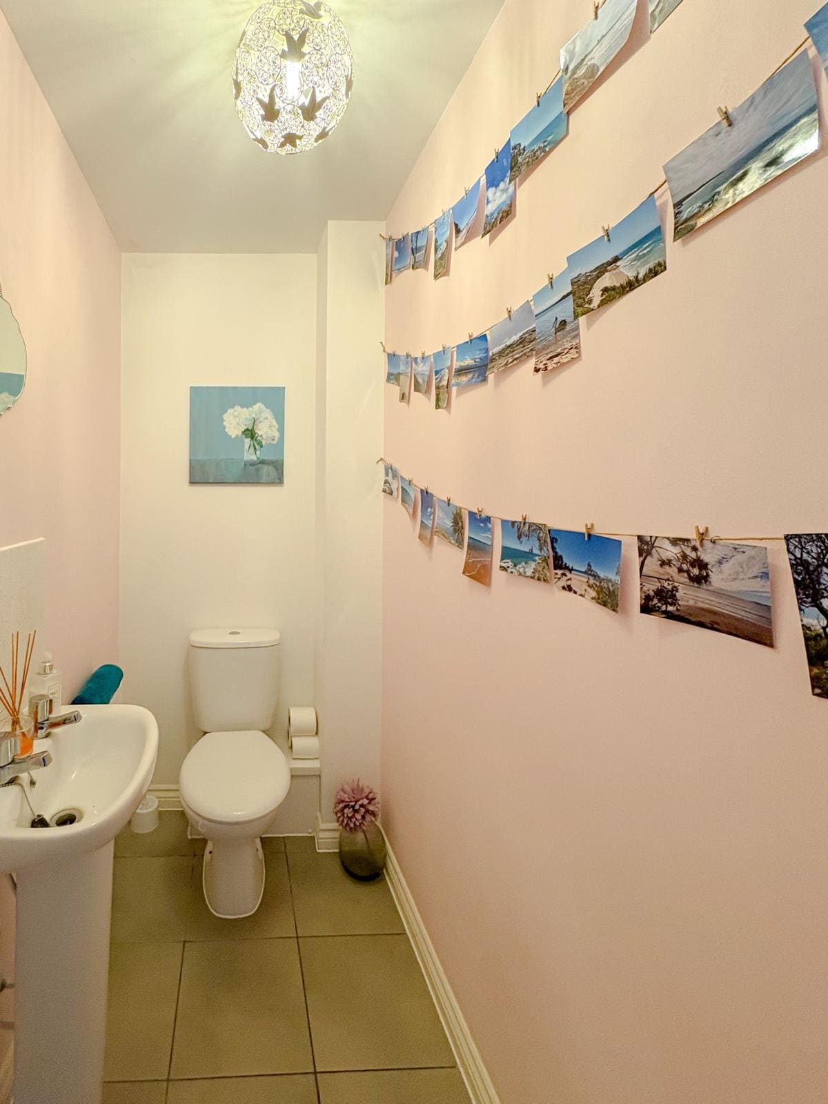 A narrow hallway is adorned with a series of photographs hung on a string, creating a visual display along the pink walls. A small white sink is positioned beside a toilet, while a decorative flower canvas adds a touch of color.