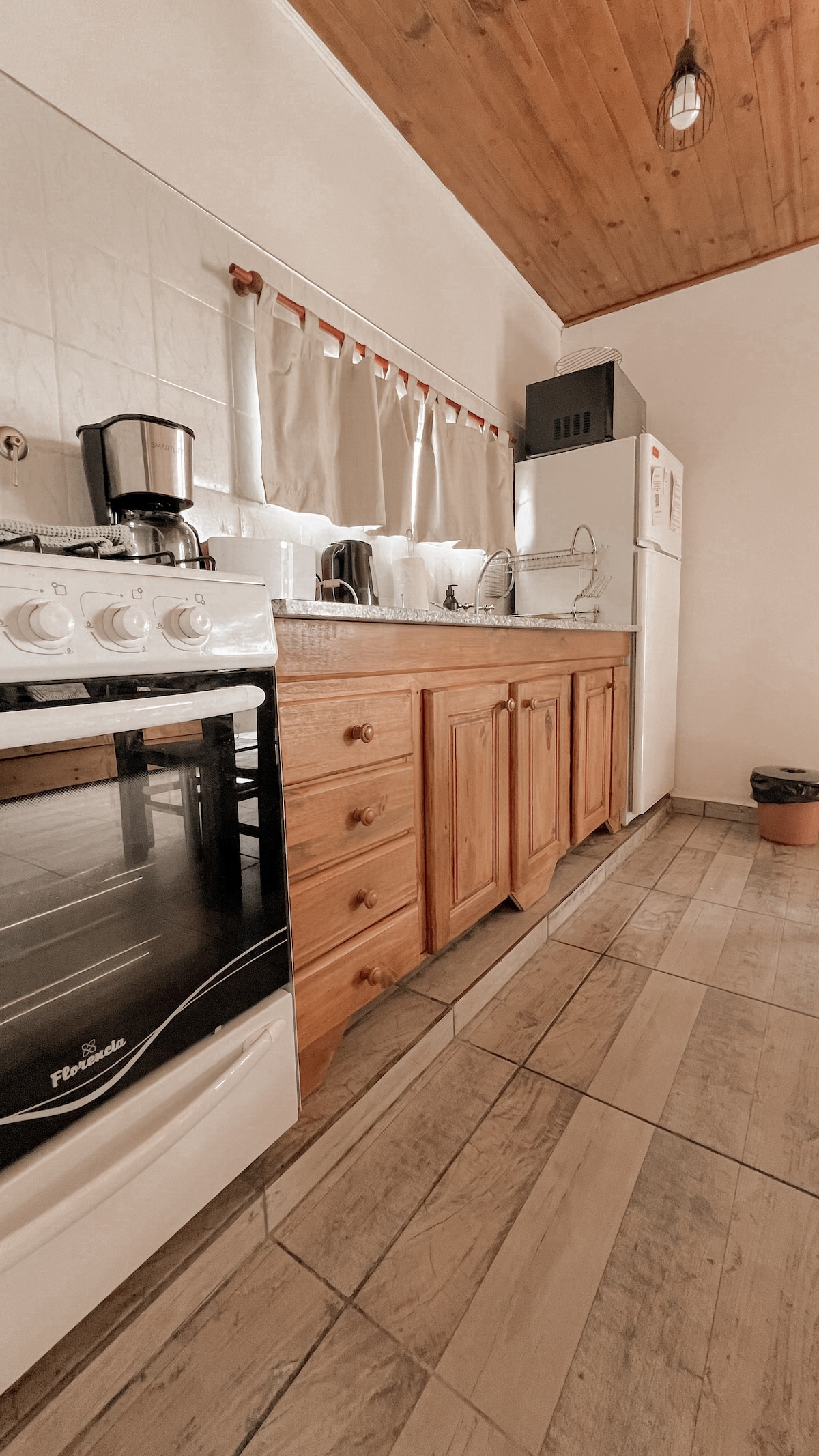 A kitchen space features wooden cabinetry and a gas stove, complemented by a counter with small appliances including a coffee maker and kettle. Light enters through a window dressed with neutral curtains, and a modern refrigerator is positioned nearby on tiled flooring.