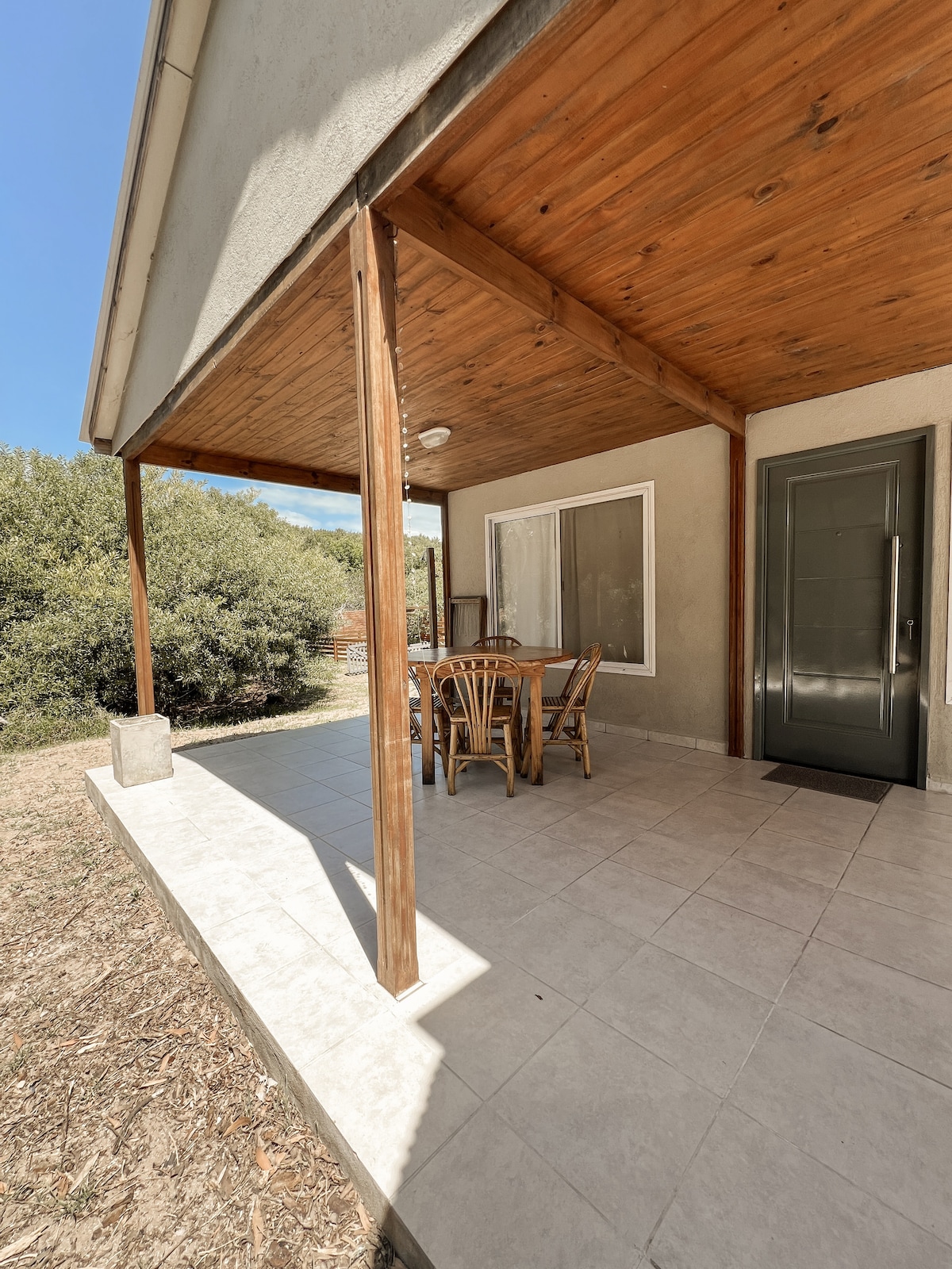 An outdoor patio features a wooden table surrounded by chairs, positioned under a sloped ceiling. A door leads into the cabin, while a well-maintained landscape with greenery is visible in the background. Natural light enhances the space under clear blue skies.
