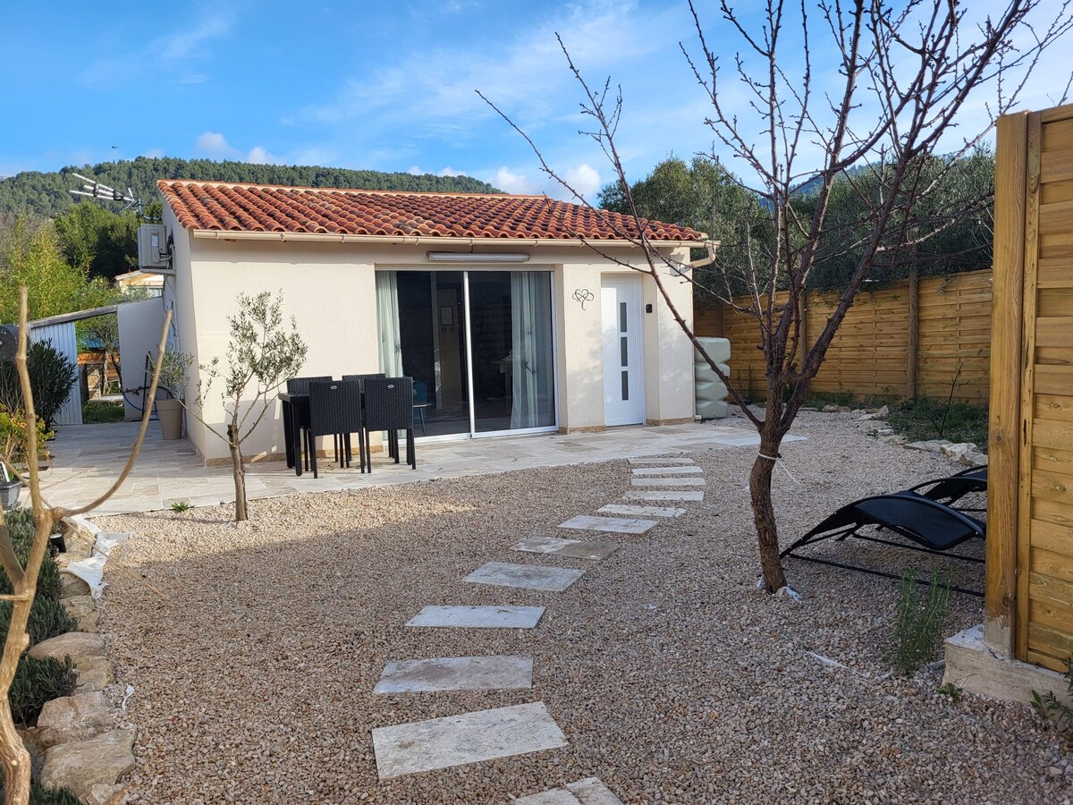 The exterior of a small house is presented, featuring a paved walkway leading to glass doors. A stone patio area includes a dining table and two lounge chairs under a tree. Surrounding greenery and a wooden fence enhance the outdoor space.