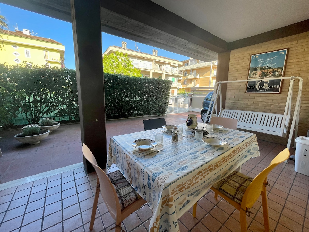 A dining area on a shaded patio features a table set for four, adorned with a tablecloth designed with various patterns. Two wooden chairs and a swing bench are visible, surrounded by greenery. Sunlight filters gently through the open space, creating a serene setting.