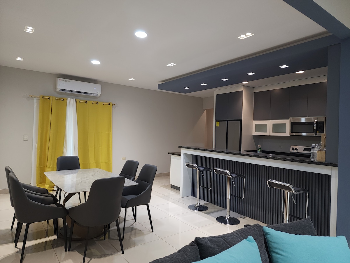 A spacious kitchen and dining area features a large round table surrounded by six modern chairs. A sleek kitchen island is equipped with bar stools, while stainless steel appliances and a contrasting grey cabinet layout are visible. Natural light filters through a window with yellow curtains.