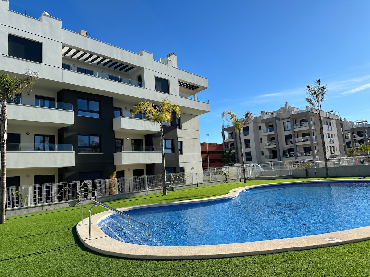 A modern building complex is depicted alongside a sparkling swimming pool, surrounded by green grass and palm trees. Clear blue skies provide a bright backdrop to the scene, enhancing the contemporary architectural design and outdoor living space.