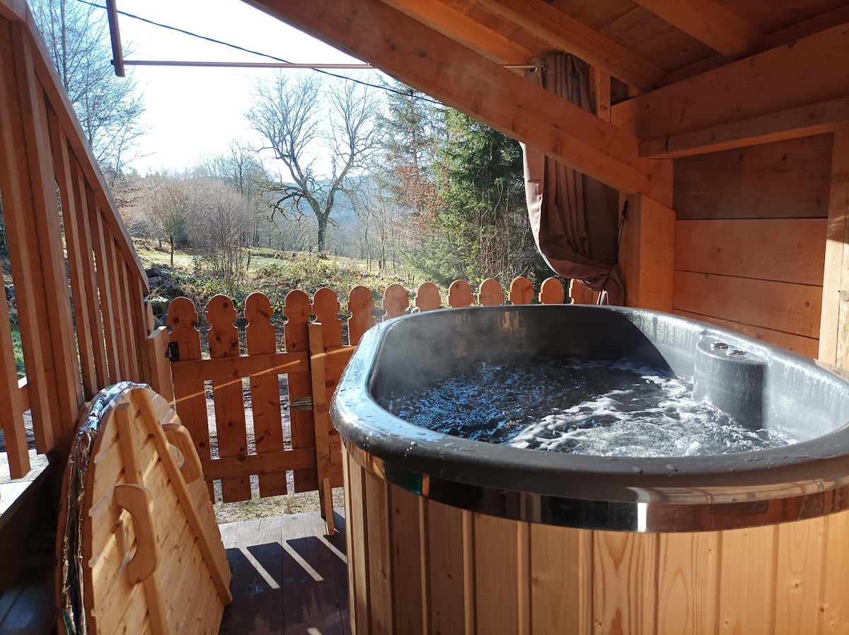 An outdoor hot tub is situated on a wooden deck, surrounded by a rustic fence. Steam rises from the water, set against a backdrop of trees and open fields under a clear blue sky. Natural light illuminates the warm wooden tones of the space.