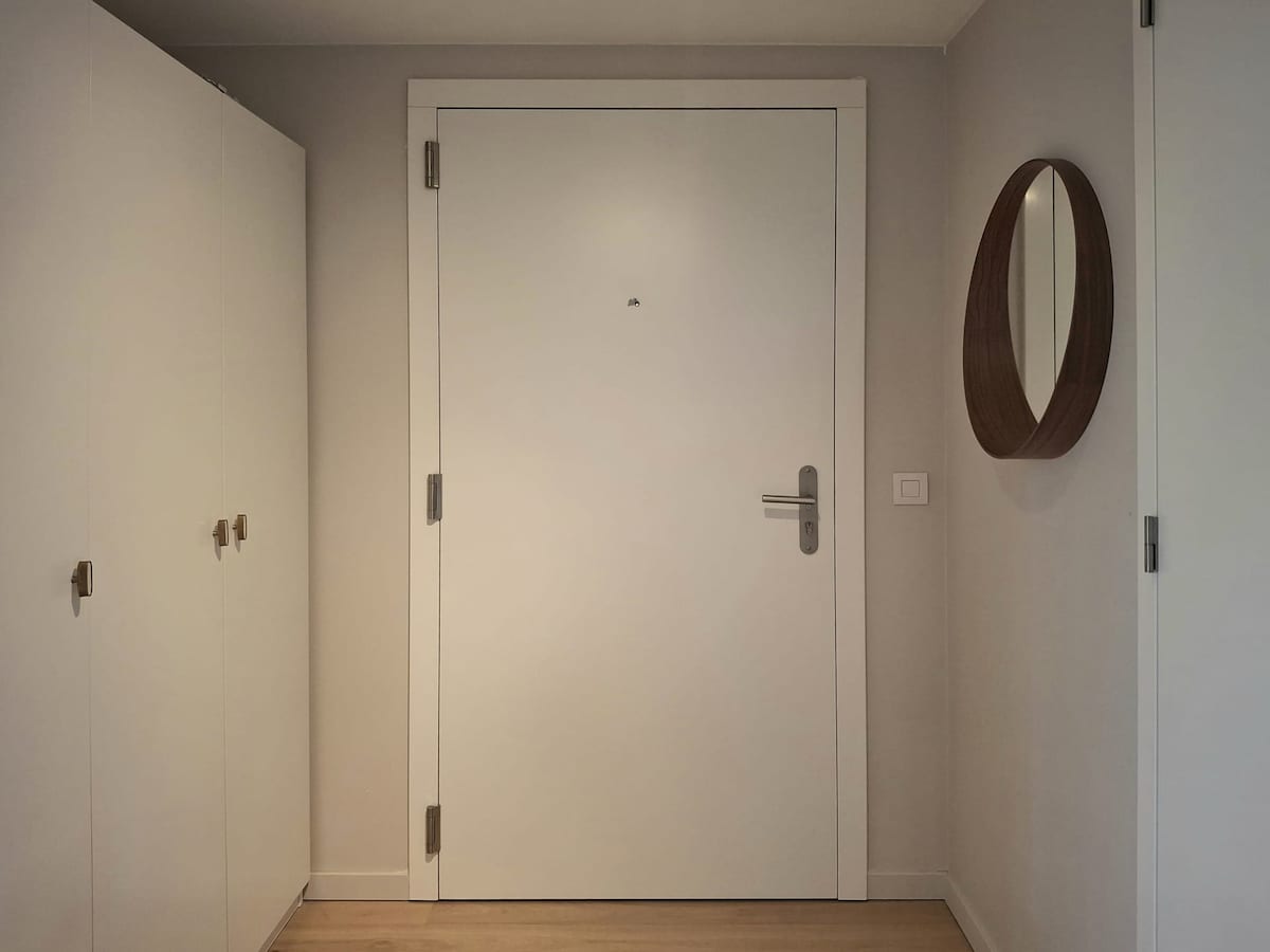 A modern entryway features a white door with a sleek handle, opposite a minimalist wardrobe with matching handles. A round mirror is hung on the wall, reflecting light and contributing to the bright ambiance. Warm wood flooring complements the neutral color palette.