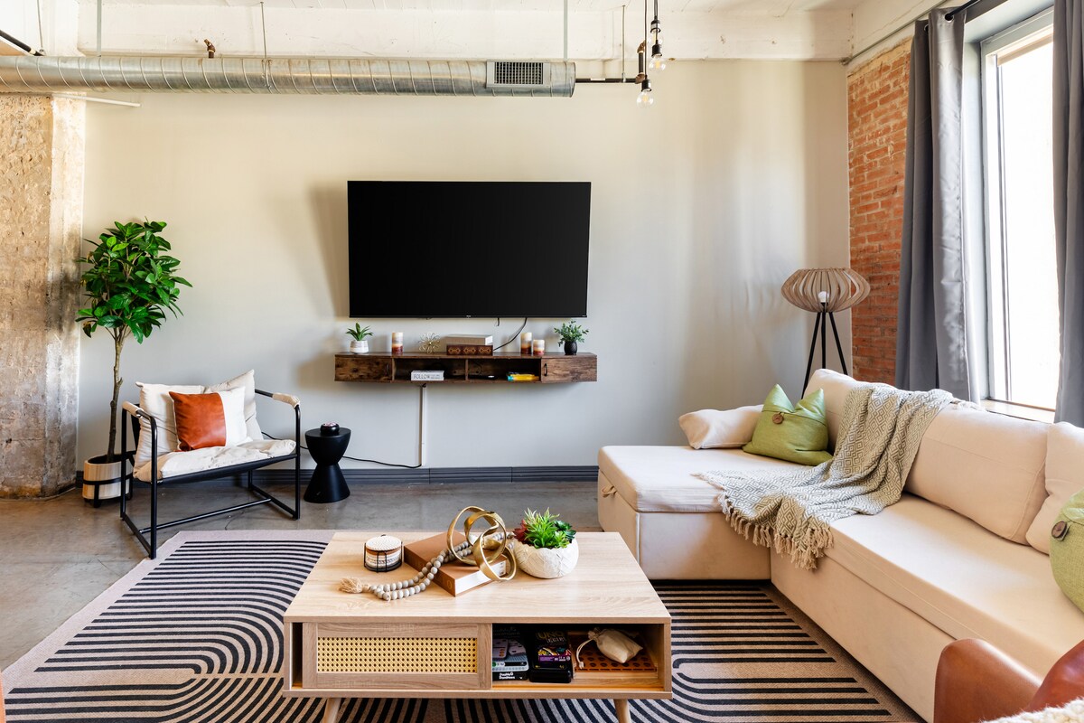 A modern living space features a light-colored sectional sofa and a woven throw draped across its arm. A coffee table with decorative items sits in the center, while a wall-mounted TV is positioned above a wooden shelf. Greenery is provided by a potted plant in the corner.