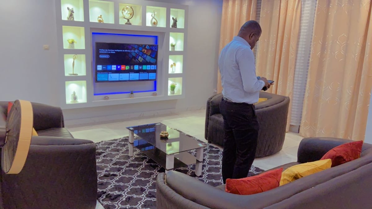 A modern living area is shown, featuring two comfortable sofas positioned around a glass coffee table. A large TV mounted on the wall displays interactive content, and decorative shelves are illuminated, showcasing various items. Soft light filters through sheer curtains, enhancing the room's inviting ambiance.