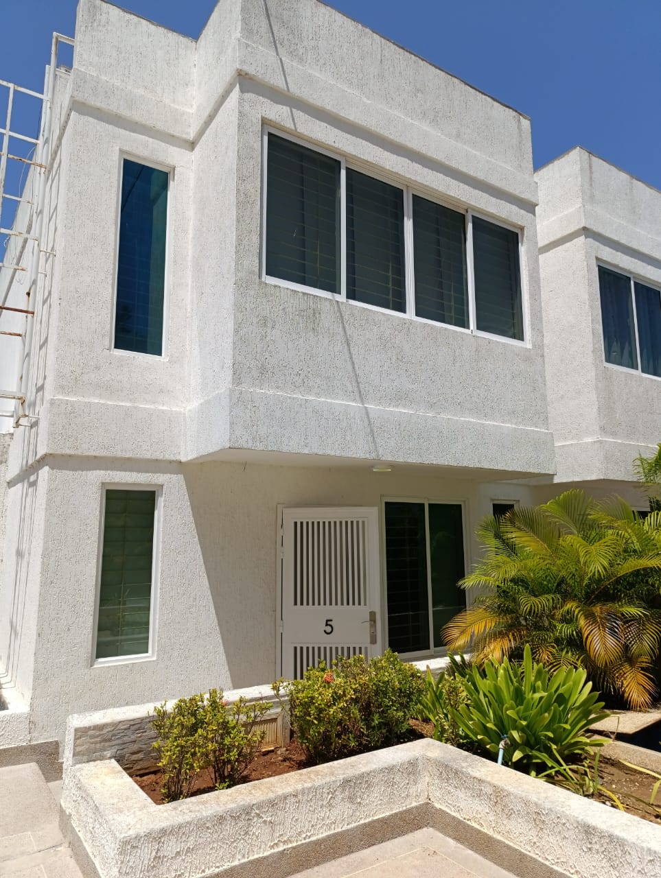 The exterior of Casa Caribe features a modern two-story design with clean lines and large windows, allowing natural light to fill the space. The bright white facade is complemented by lush greenery and a small garden area in front, creating a welcoming entrance.