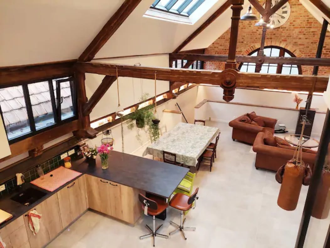 An open kitchen space features wooden beams and modern fixtures, complemented by a central black bar counter with stools. A dining table is set nearby, surrounded by natural light from large windows, while a comfortable seating area is visible in the background.