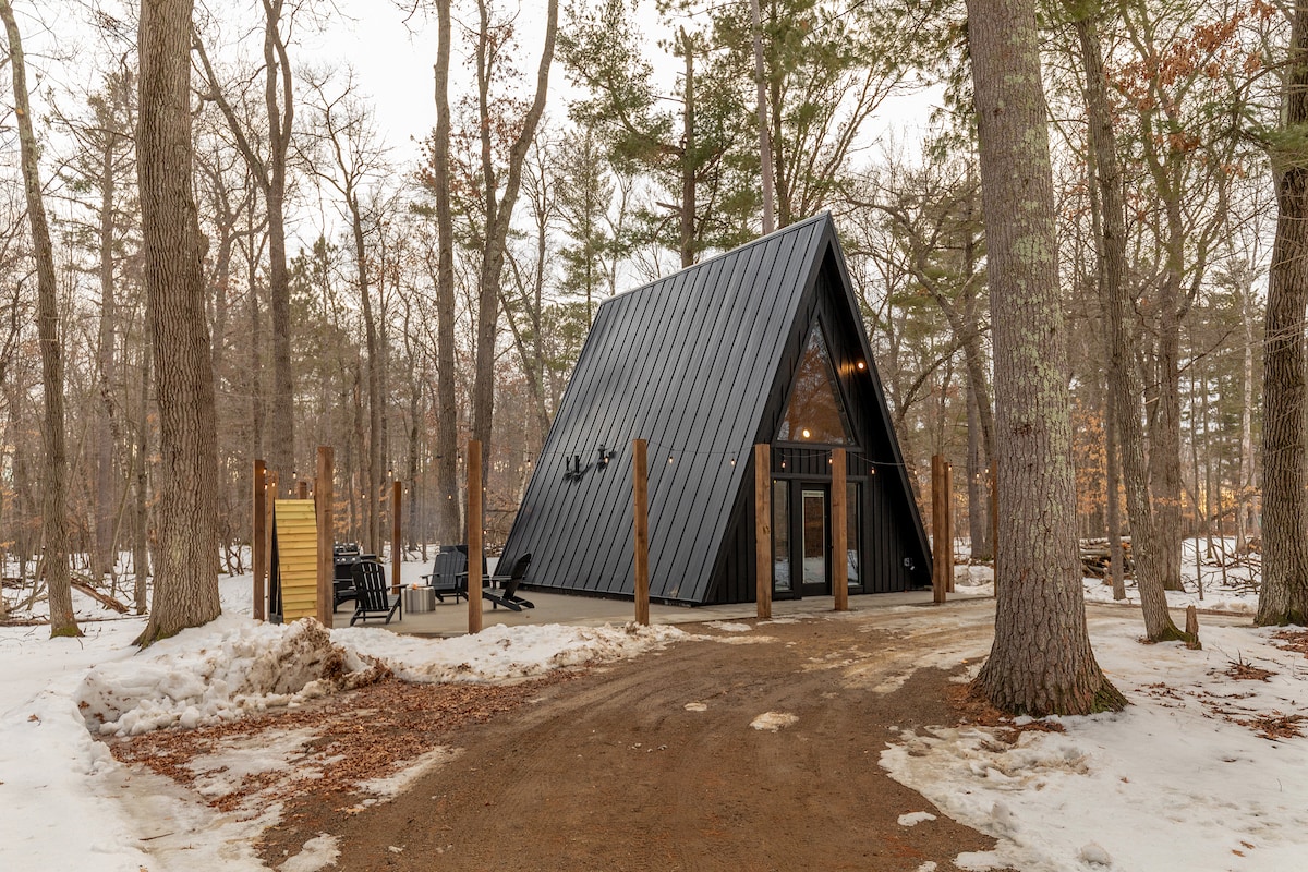 The A-frame cabin is nestled among tall trees, featuring a striking black exterior with a metal roof. A gravel path leads to the entrance, bordered by snow-dusted grounds. Outdoor seating and a fire pit can be seen, providing a tranquil setting for gatherings.