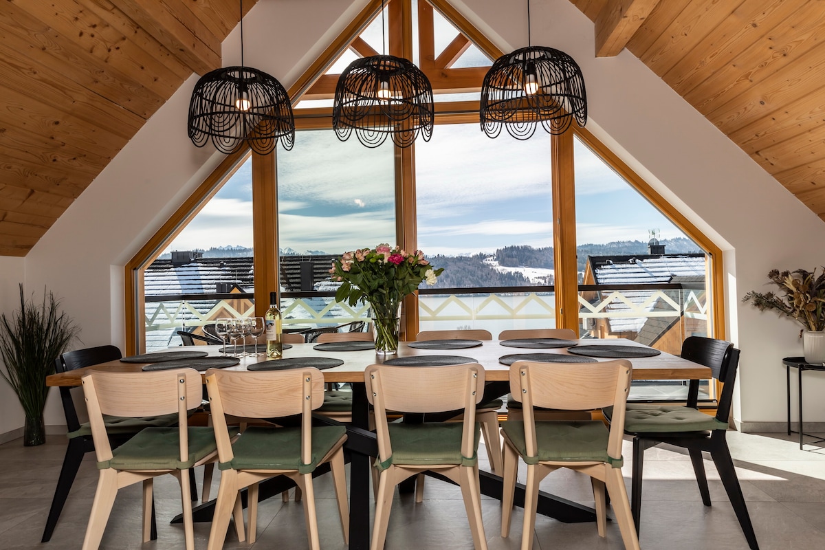 A spacious dining area features a large table surrounded by ten chairs, with a bouquet of flowers at its center. Large triangular windows flood the space with natural light, offering scenic views of the surrounding landscape and mountains.