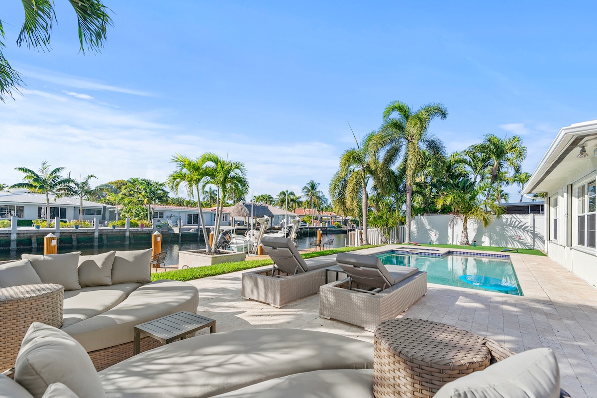 A serene outdoor space features a private pool surrounded by lounge chairs and a sectional sofa. Lush palm trees provide shade, while the scenic waterway enhances the retreat's tranquil atmosphere. The area is designed for relaxation, with ample seating for guests to enjoy the waterfront view.