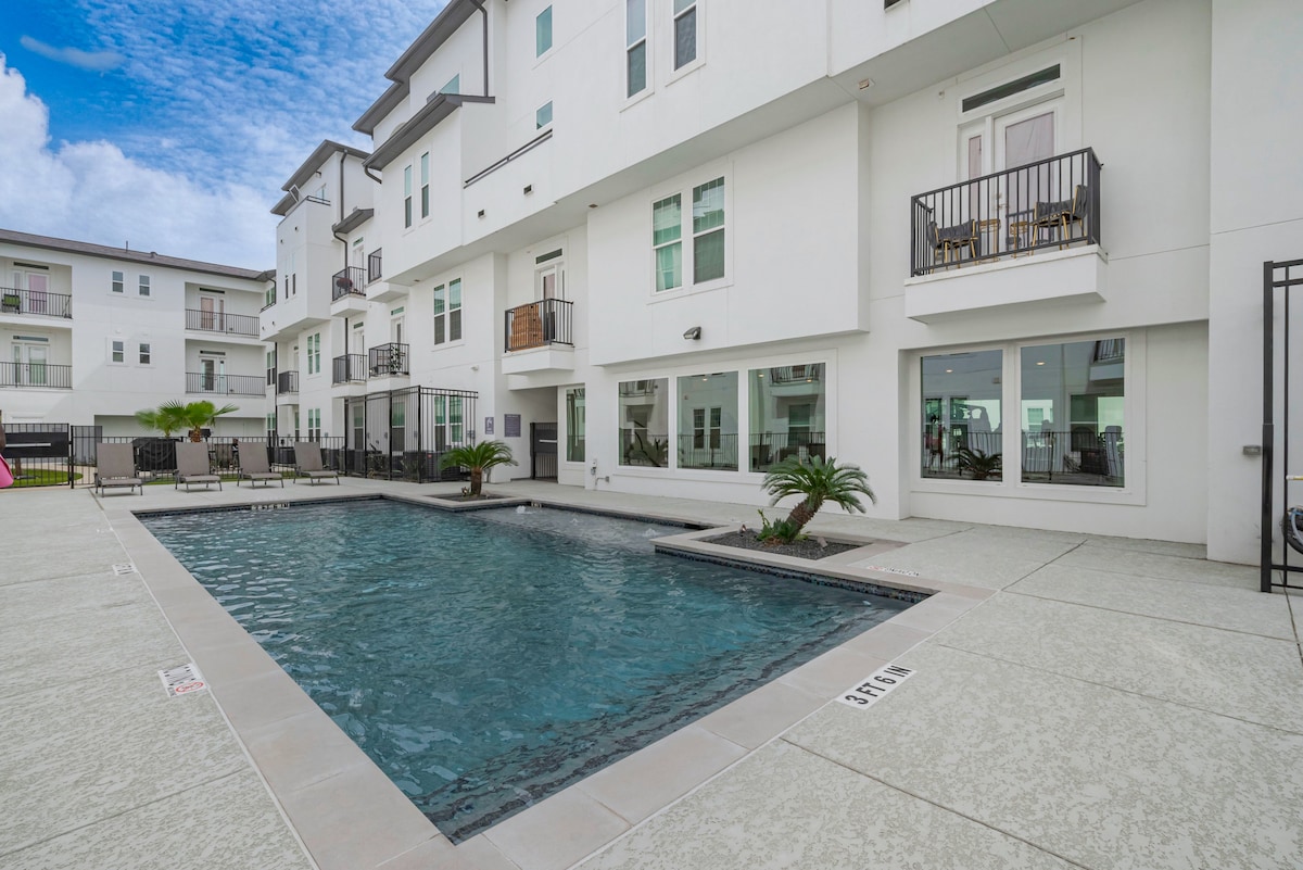 A modern outdoor area features a clear swimming pool surrounded by a textured patio. Sun loungers are arranged around the pool, providing comfortable seating options. Balconies from nearby apartments overlook the pool area, with a landscaped palm adding a touch of green.