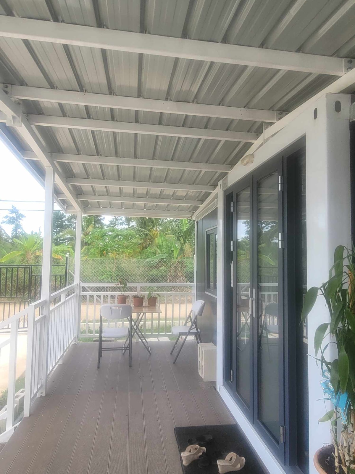 A shaded porch area features a metal roof and a wooden floor. Two chairs and a small table are positioned for seating, surrounded by greenery and a view of the outdoor space. Natural light filters in through large glass doors.