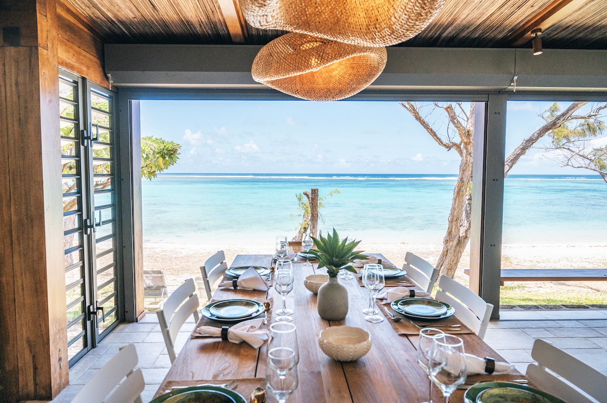 An inviting dining area is highlighted by a large wooden table set with utensils and glassware. Oversized light fixtures hang above, while wide glass doors offer a view of the tranquil beach and ocean, enhancing the connection to the outdoor environment.