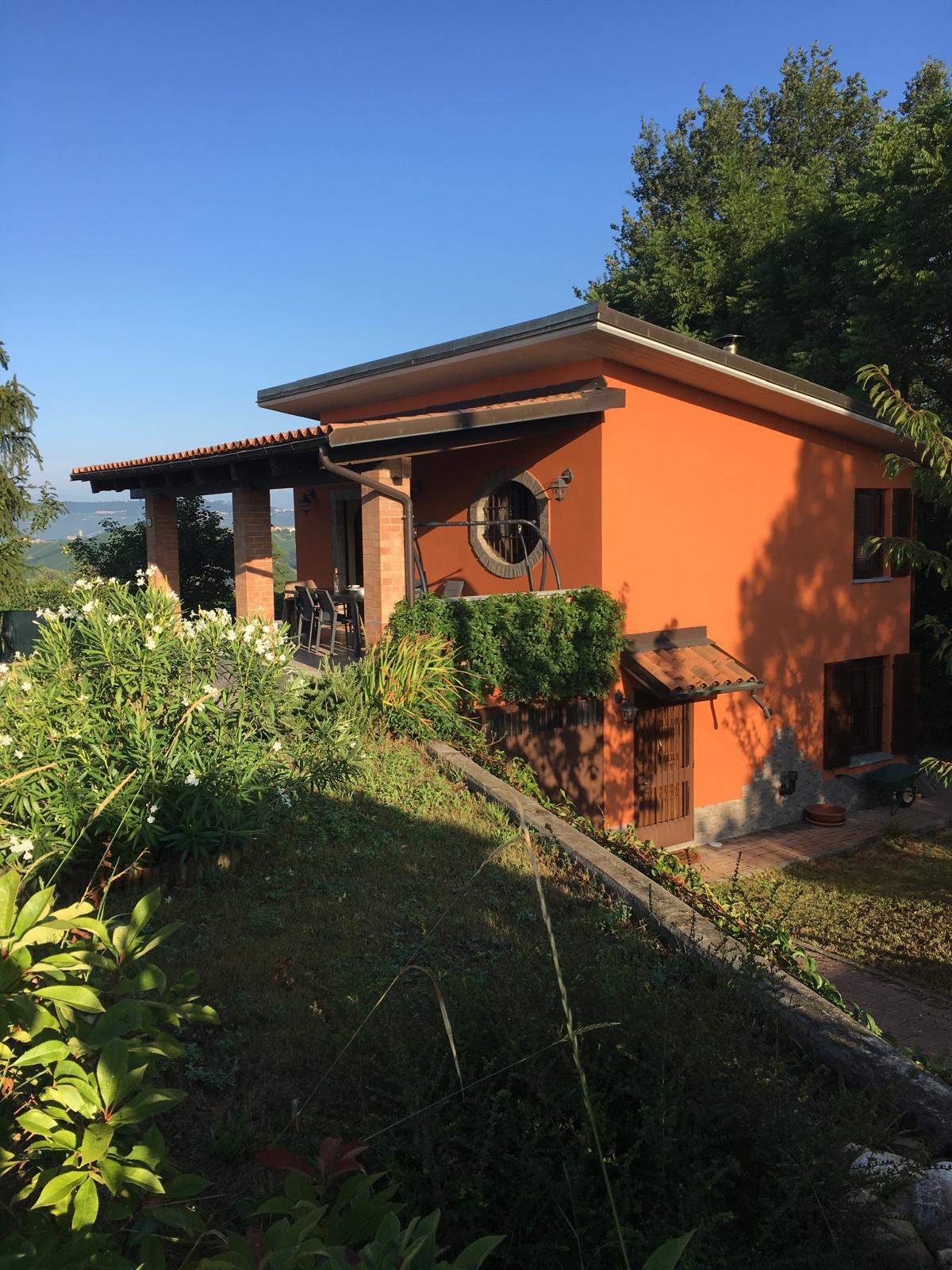 The exterior of the vibrant orange house is surrounded by lush greenery, showcasing a blend of flowers and plants. A spacious terrace is visible, equipped with a table and chairs, inviting relaxation. The structure sits on a gentle slope, framed by trees and the clear blue sky.