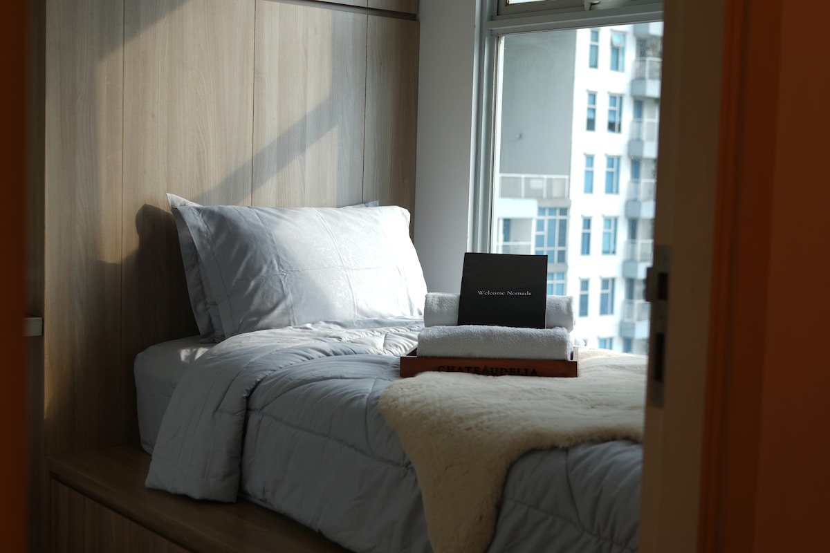 A cozy sleeping area showcases a spacious bed dressed in light grey linens. A soft cream throw is casually placed on the edge. Natural light filters through the window, revealing a view of the city skyline.